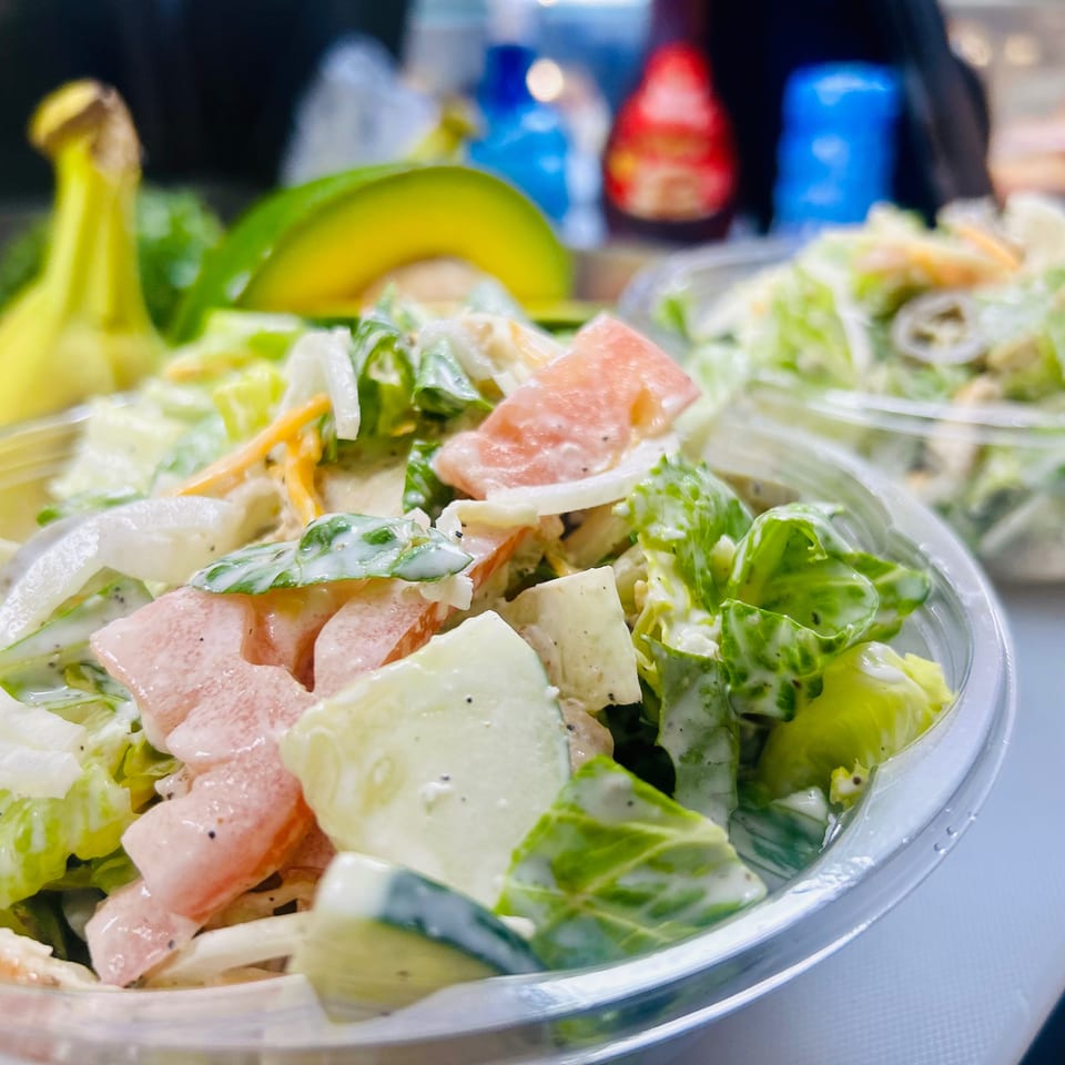 Two plastic containers of fresh salad with lettuce, tomatoes, and dressing, with fruit in the background.
