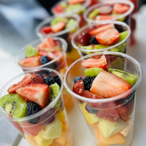 Rows of clear plastic cups filled with colorful fresh fruit salad, including strawberries, kiwi, and berries.
