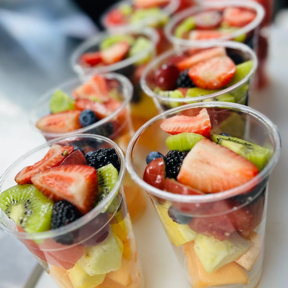 Rows of clear plastic cups filled with colorful fresh fruit salad, including strawberries, kiwi, and berries.