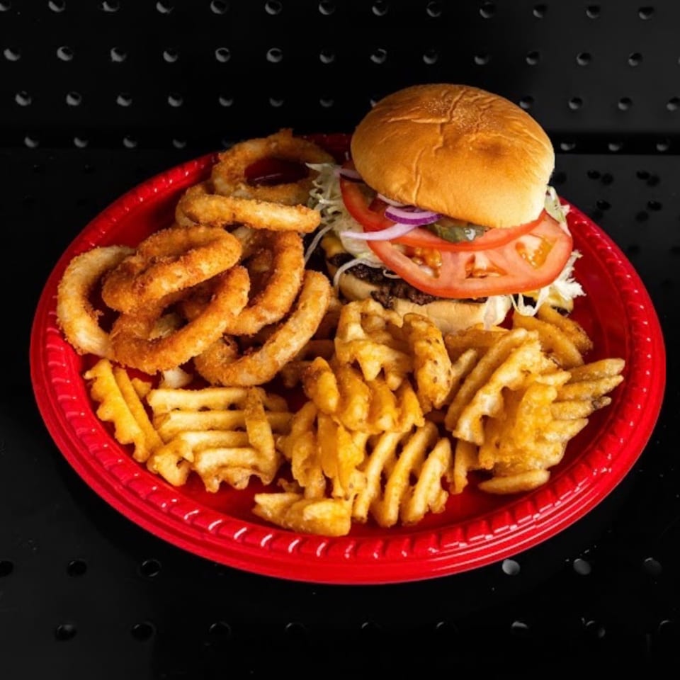 A cheeseburger with lettuce, tomato, onion, and pickles, served with waffle fries and onion rings on a red plate.